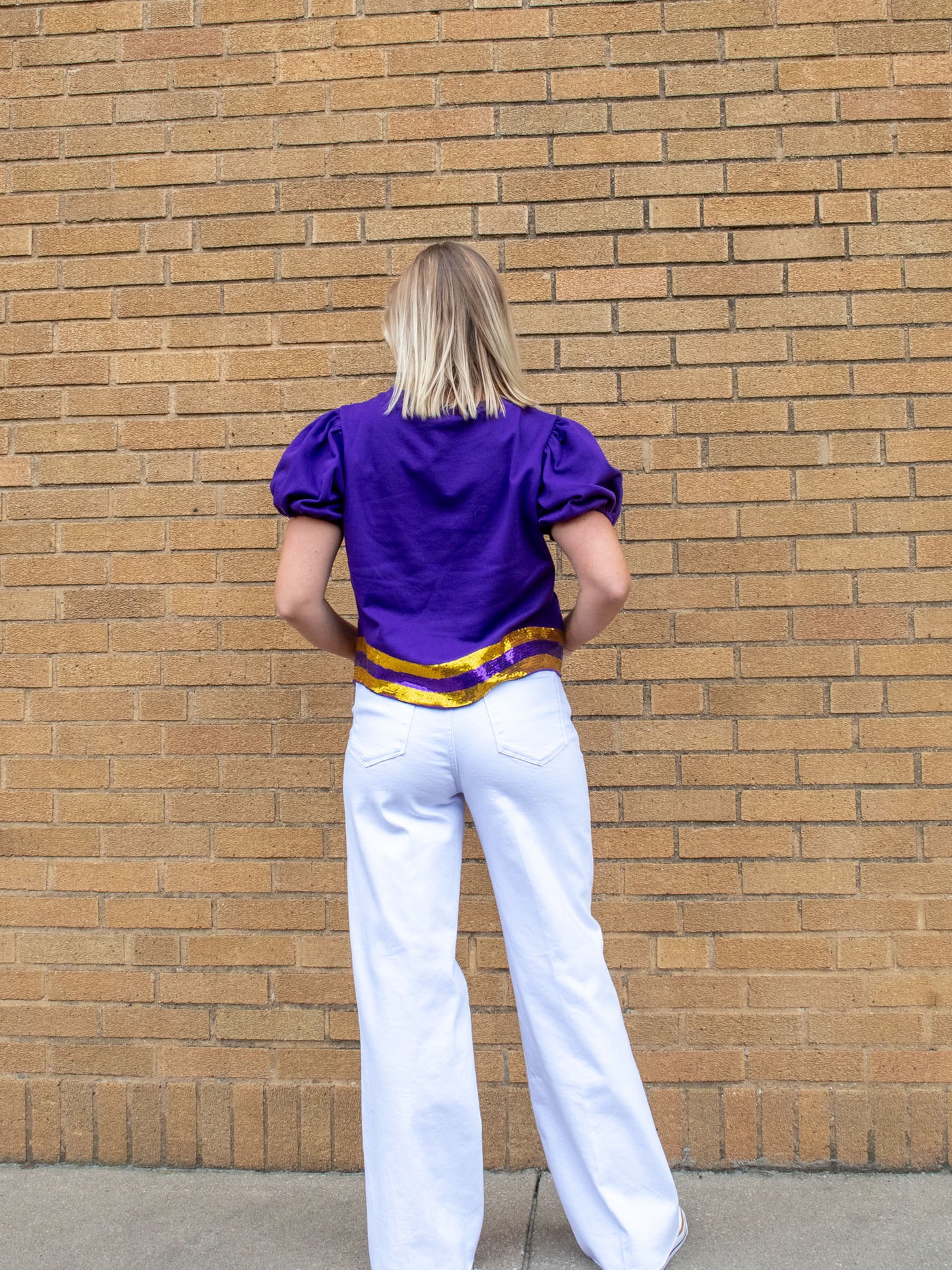 A model wearing a purple top that says touchdown and has purple and gold sequins. The model has it paired with a white jean.
