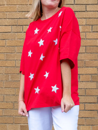 A model wearing a red oversized tee with white sparkly stars on it. The model has it paired with a white jean.