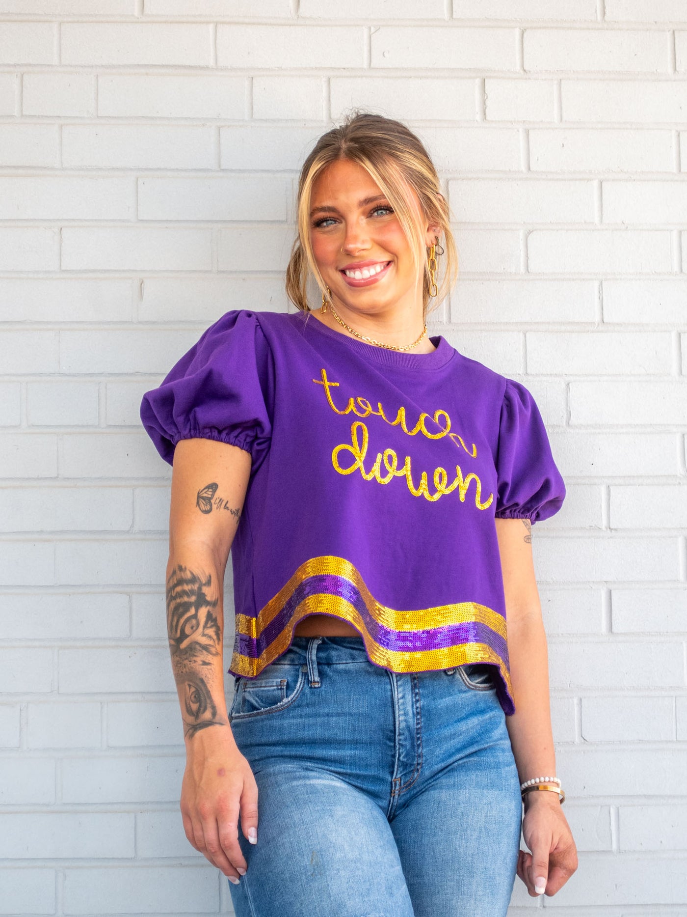 A model wearing a purple tee with a gold and purple sequin wavy hem and the phrase "touch down" on it. She has it paired with a pair of light wash jeans.
