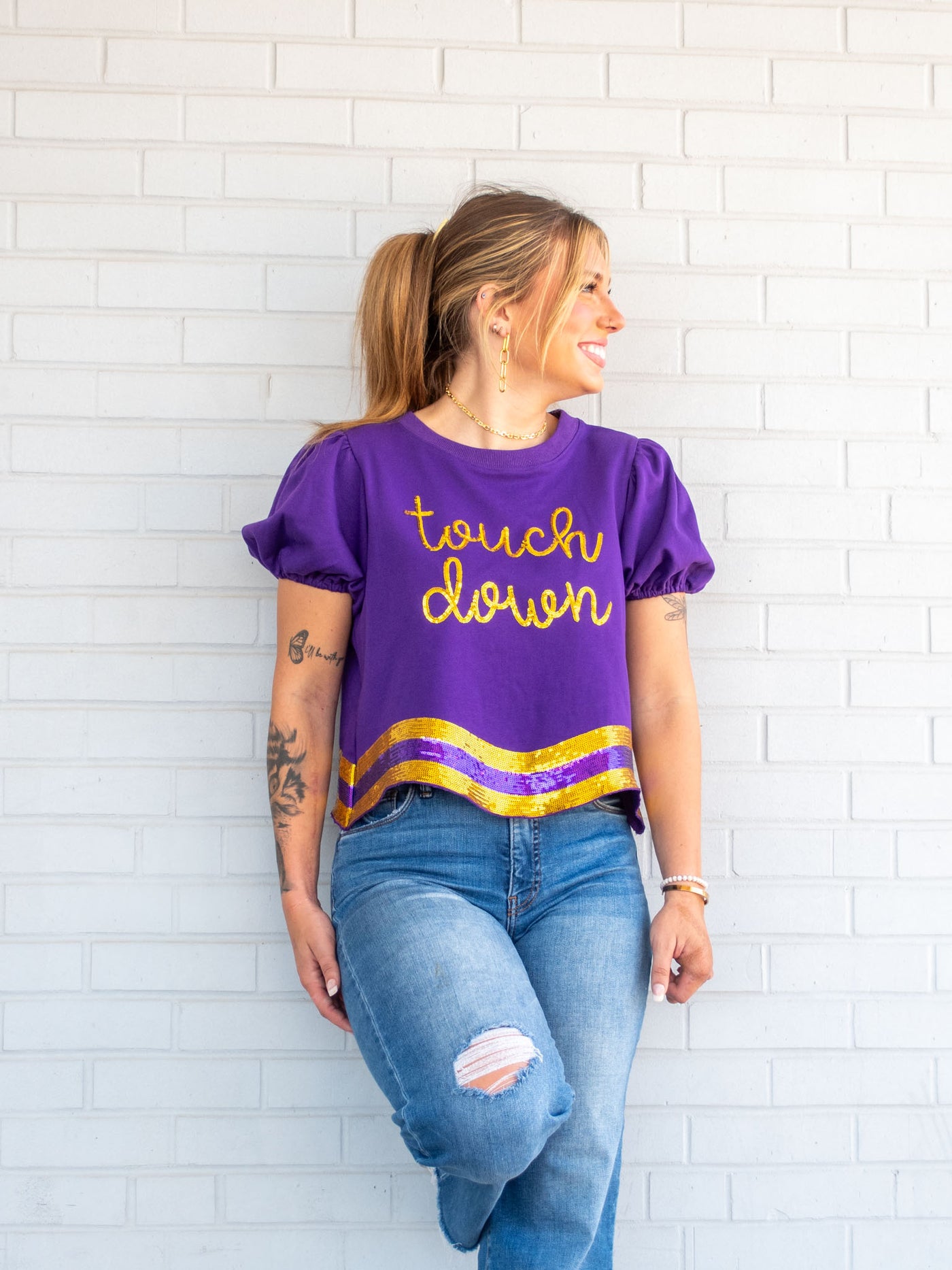 A model wearing a purple tee with a gold and purple sequin wavy hem and the phrase "touch down" on it. She has it paired with a pair of light wash jeans.