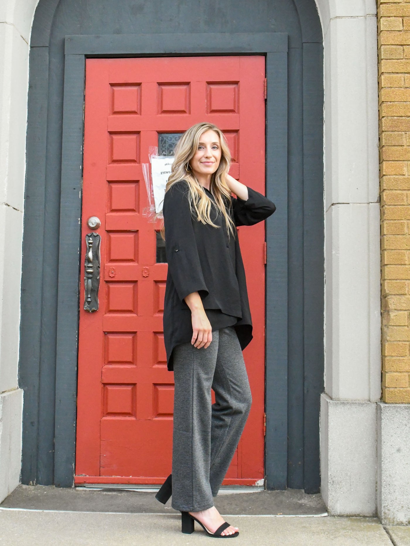 A model wearing a black airflow top with a v-neckline and adjustable sleeves. The model has it paired with a wide leg trouser and black heels.