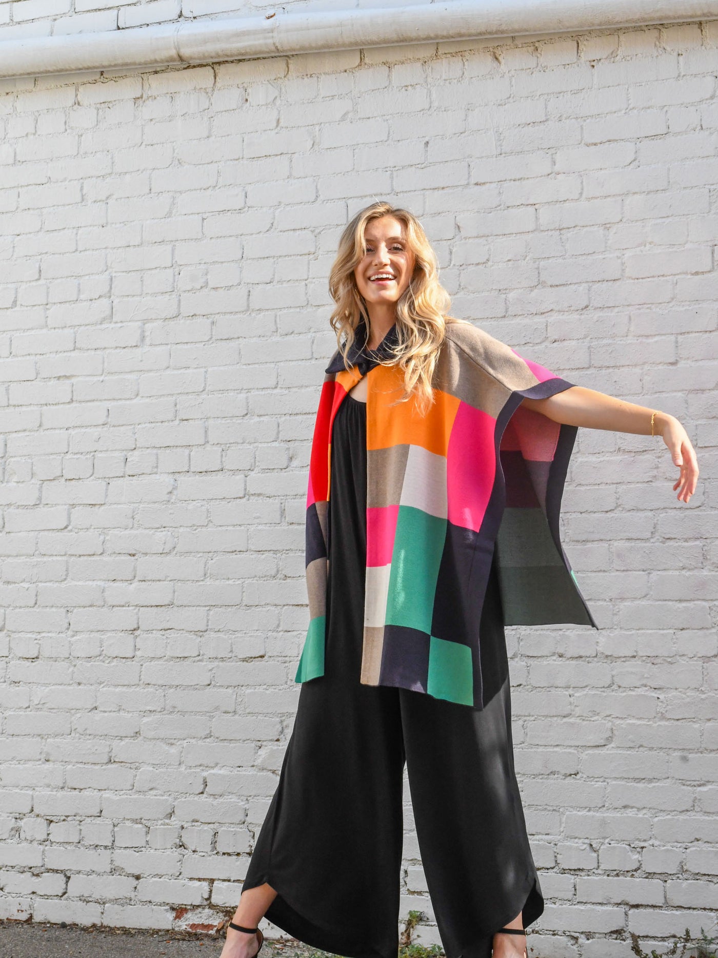 A model wearing a purple, red, orange, pink, green, grey and navy color blocked poncho with a collar. The model is wearing it on top of a black jumpsuit.