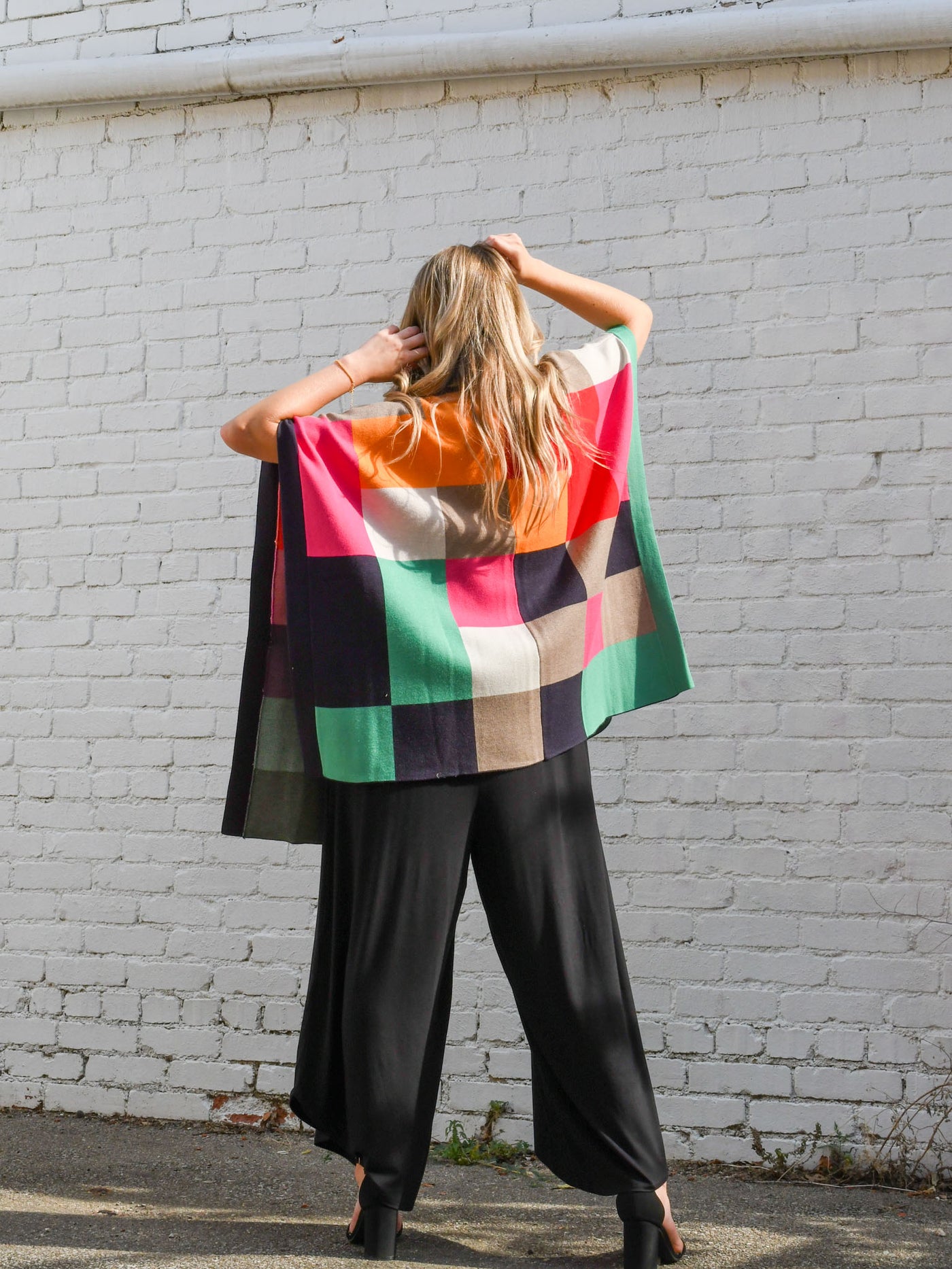 A model wearing a purple, red, orange, pink, green, grey and navy color blocked poncho with a collar. The model is wearing it on top of a black jumpsuit and black heels.