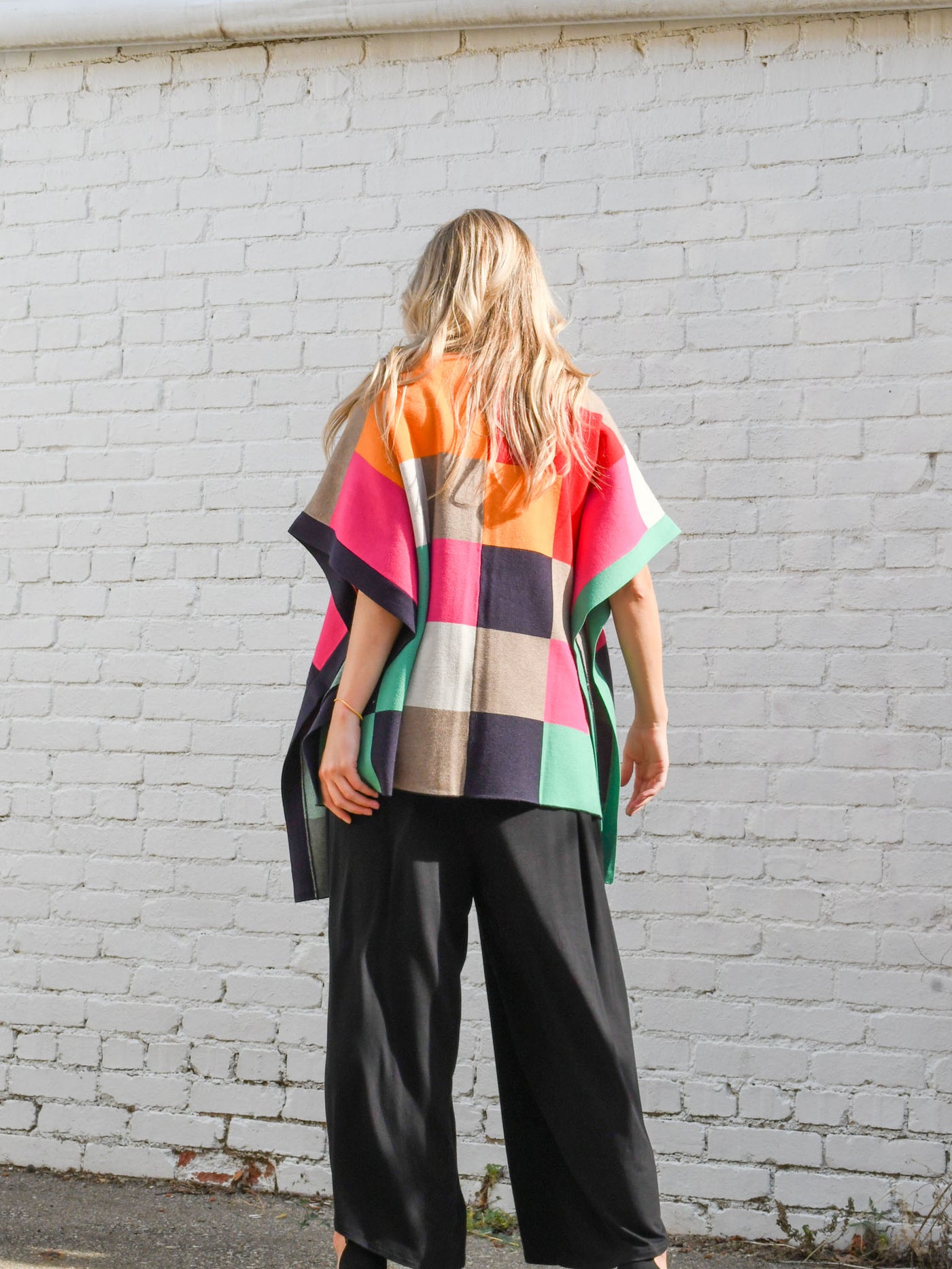 A model wearing a purple, red, orange, pink, green, grey and navy color blocked poncho with a collar. The model is wearing it on top of a black jumpsuit.