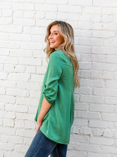 A model wearing a green airflow blouse pinned up sleeves and a v-neckline with collar. She has it paired with dark wash jeans.