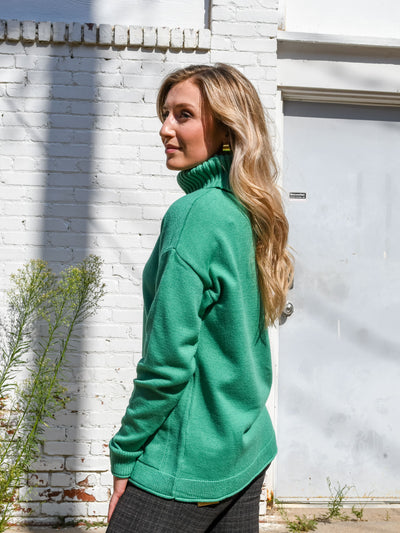 A model wearing a green turtleneck sweater with seam details. The model has it paired with a black and brown plaid legging.