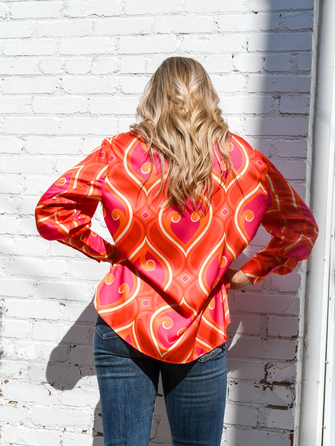 A model wearing a pink and orange printed silk top with a v-neckline. The model has it paired with a pair of dark wash jeans.