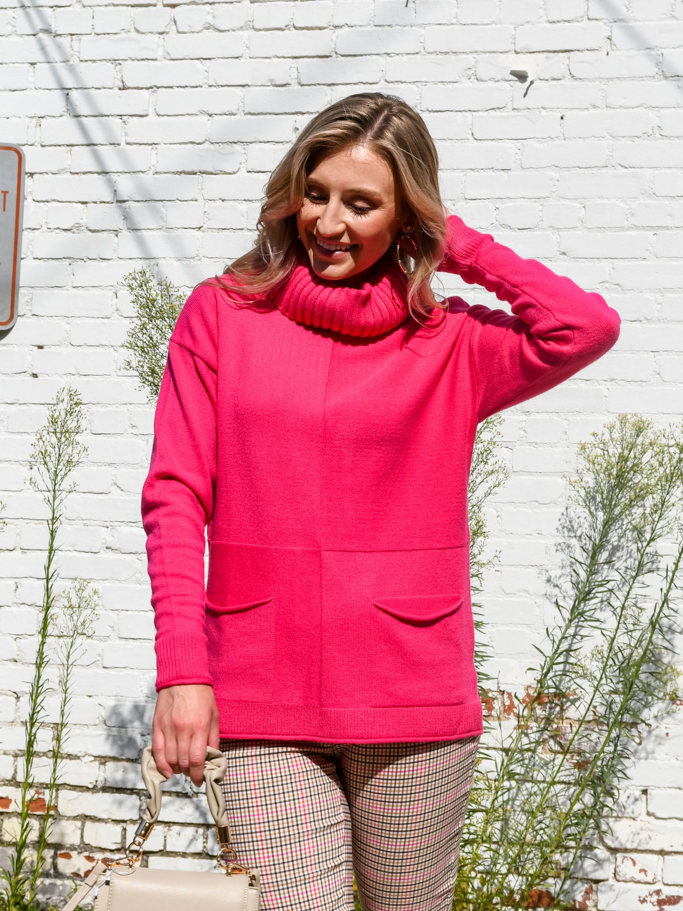 A model wearing a hot pink turtleneck sweater with seam details. The model has it paired with a pink and brown plaid trouser.