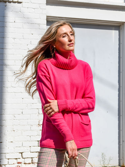 A model wearing a hot pink turtleneck sweater with seam details. The model has it paired with a pink and brown plaid trouser.