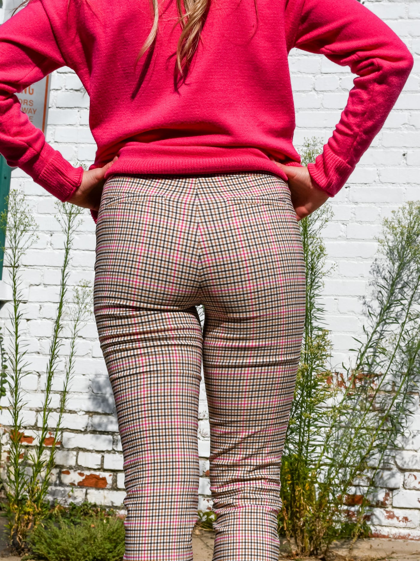 A model wearing a pair of pink plaid ankle length dress pants. The model has them paired with a pink turtleneck sweater, a beige purse and brown sandals.