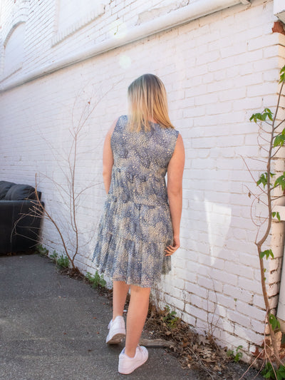 A model wearing a blue sleeveless mesh tiered shift dress that has faux buttons and a floral pattern. She has it on with white sneakers.