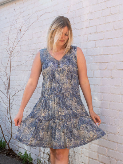 A model wearing a blue sleeveless mesh tiered shift dress that has faux buttons and a floral pattern. She has it on with white sneakers.