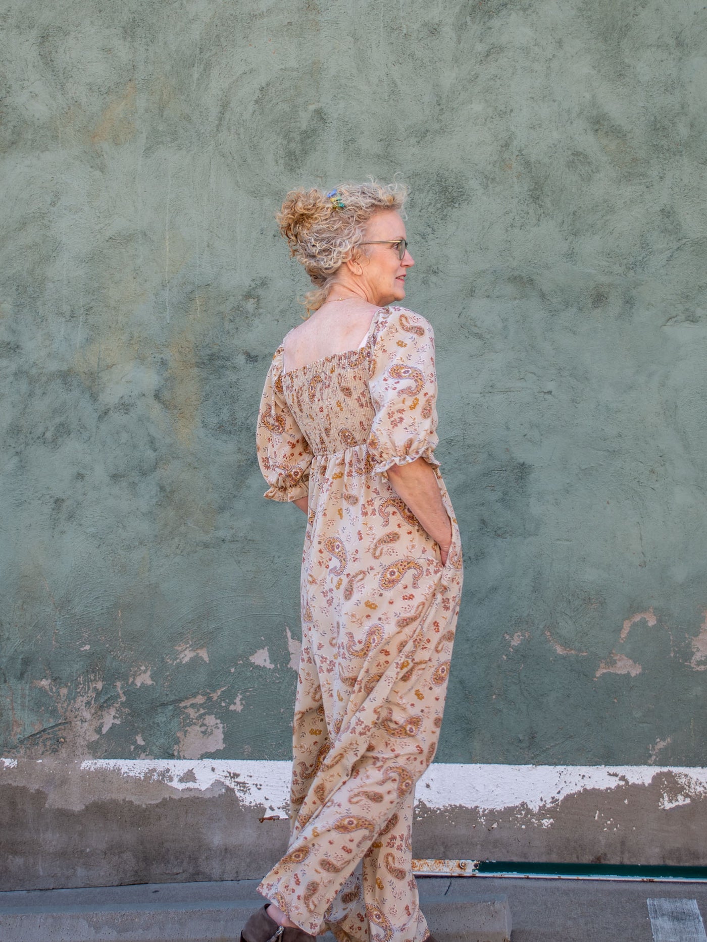 A model wearing a tan, paisley printed jumpsuit with puff sleeves and a square neckline. The model has it paired with a gray bootie.