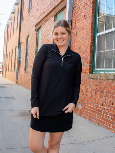 A model wearing a black quarter zip active mock neck top with a black skort and white sneakers.