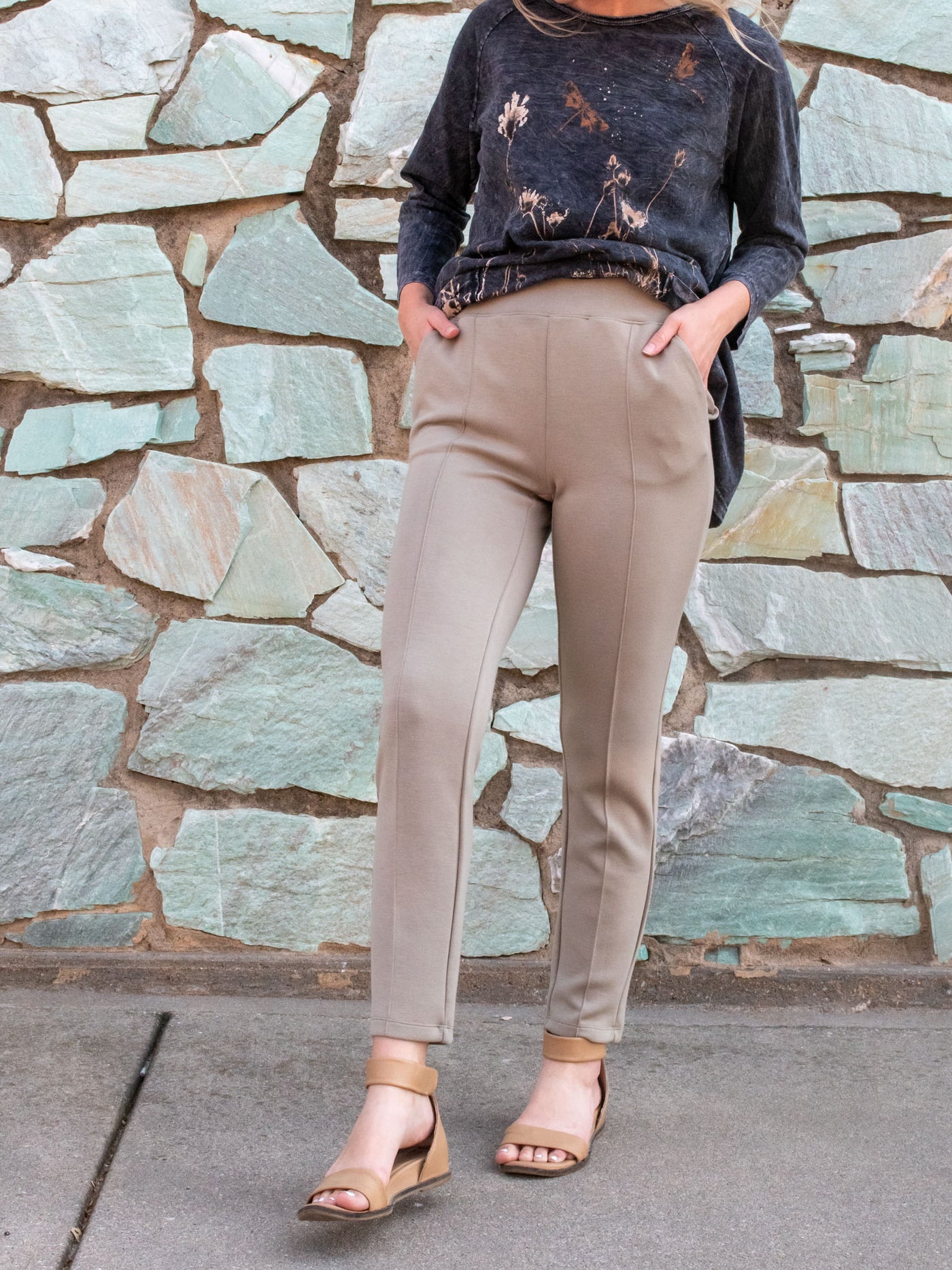 A model wearing a pair of tan scuba knit, ankle length pants. The model has it paired with a grey top and nude sandals.