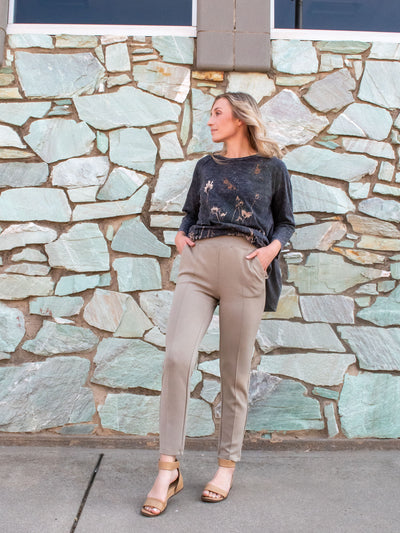 A model wearing a pair of tan scuba knit, ankle length pants. The model has it paired with a grey top and nude sandals.