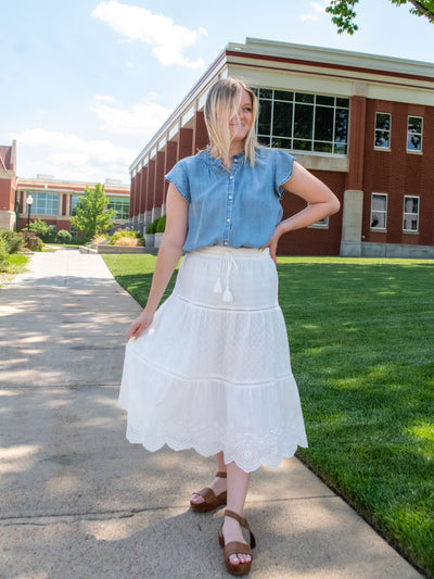 A model wearing a white maxi embroidery skirt with scalloped edge. She has it on with a blue blouse and sandals.