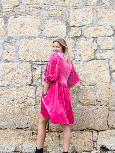 A model wearing a fuchsia velvet dress with a dolman sleeve and pockets. The model has it paired with a black bootie.