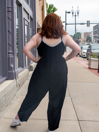A model wearing a wide leg, black jumpsuit. She has it layered over a light gray tank top and white sneakers.