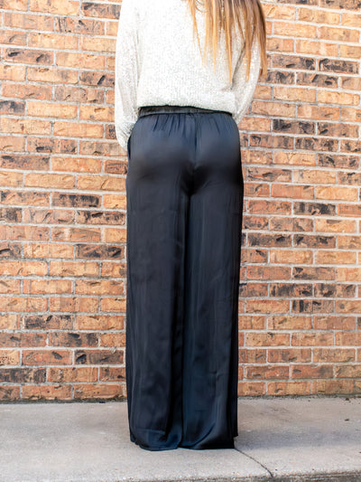 A model wearing a pair of silk, black flowy pants. The model has them paired a white sparkly top and black booties.
