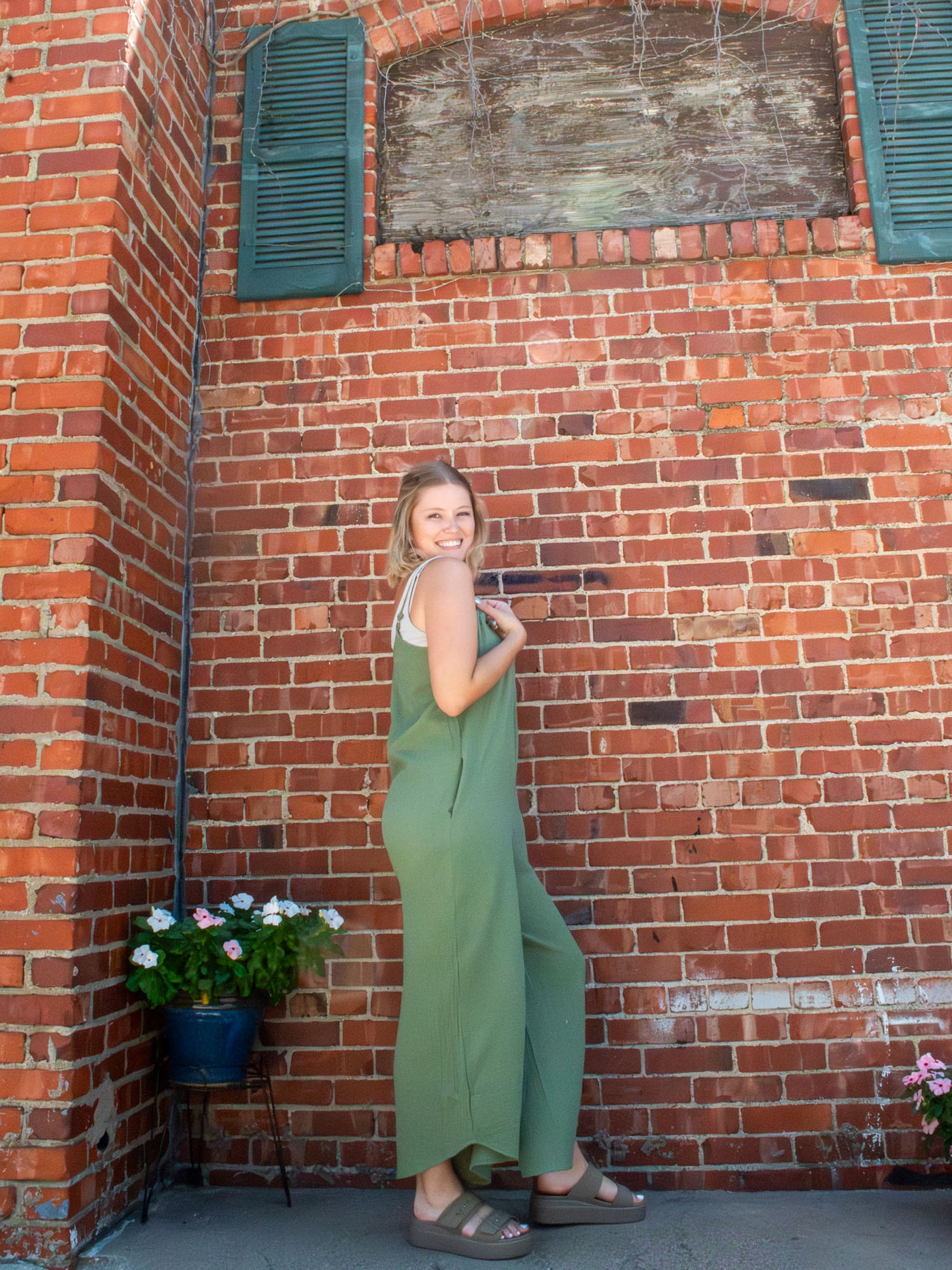A model wearing an olive green, gauze jumpsuit. The model paired it with a beige tank underneath and beige platform sandals.