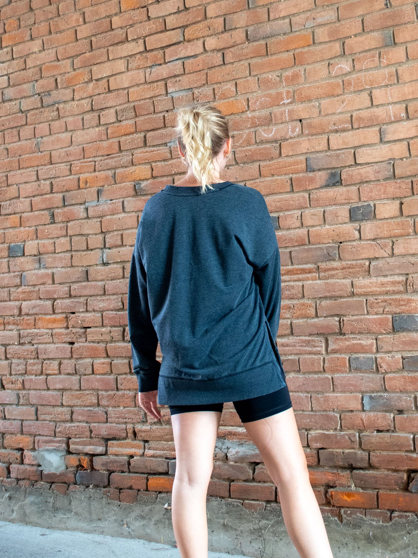 A model wearing a gray v-neck sweatshirt with an oversized look and thick hemline. The model has it paired with black biker shorts.