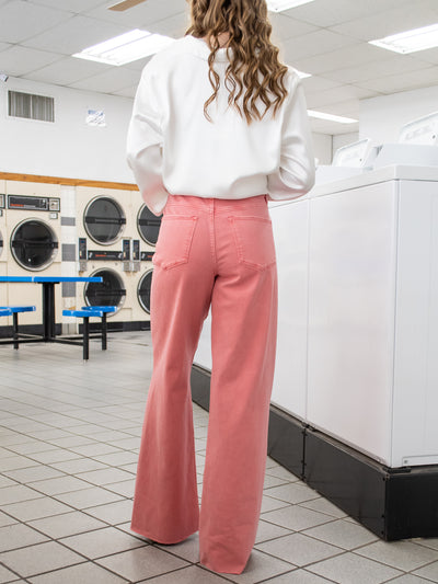 A model wearing pink straight leg jeans with an unfinished hem. She has it paired with a white silky blouse and white boots.