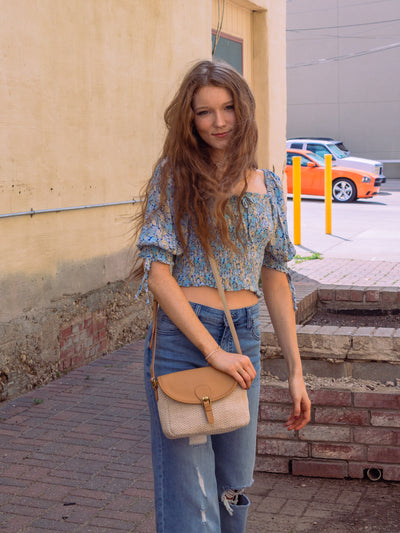 A model wearing a blue puff sleeve crop top with a smocked bodice. It has yellow and blue flowers on it. The model has it paired with a tan and white crossbody bag, blue denim wide leg jeans, and a pair of black platform sandals.
