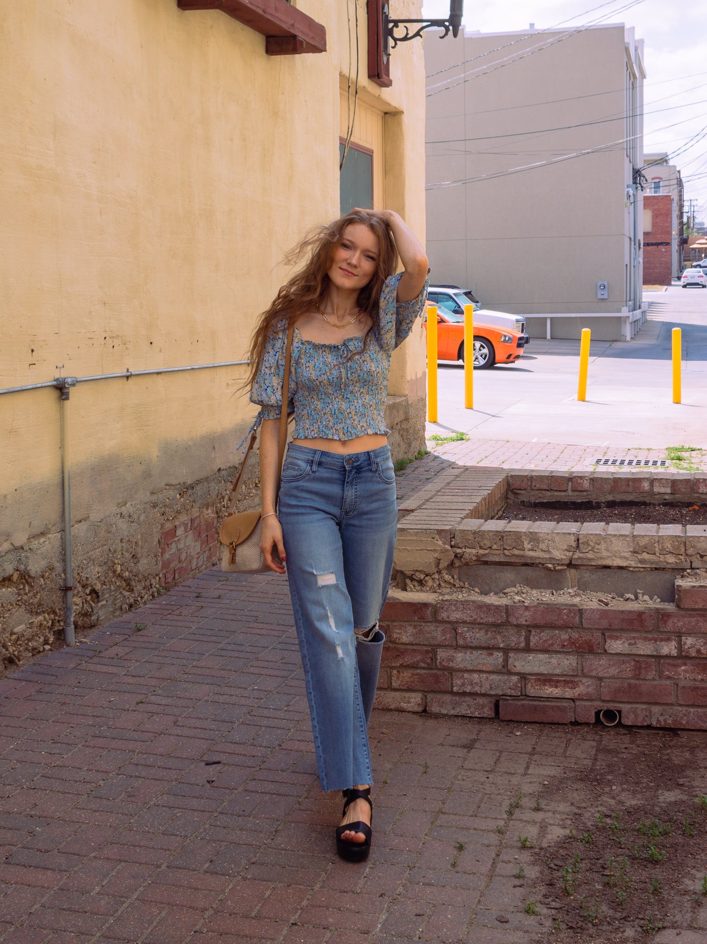 A model wearing a blue puff sleeve crop top with a smocked bodice. It has yellow and blue flowers on it. The model has it paired with a tan and white crossbody bag, blue denim wide leg jeans, and a pair of black platform sandals.
