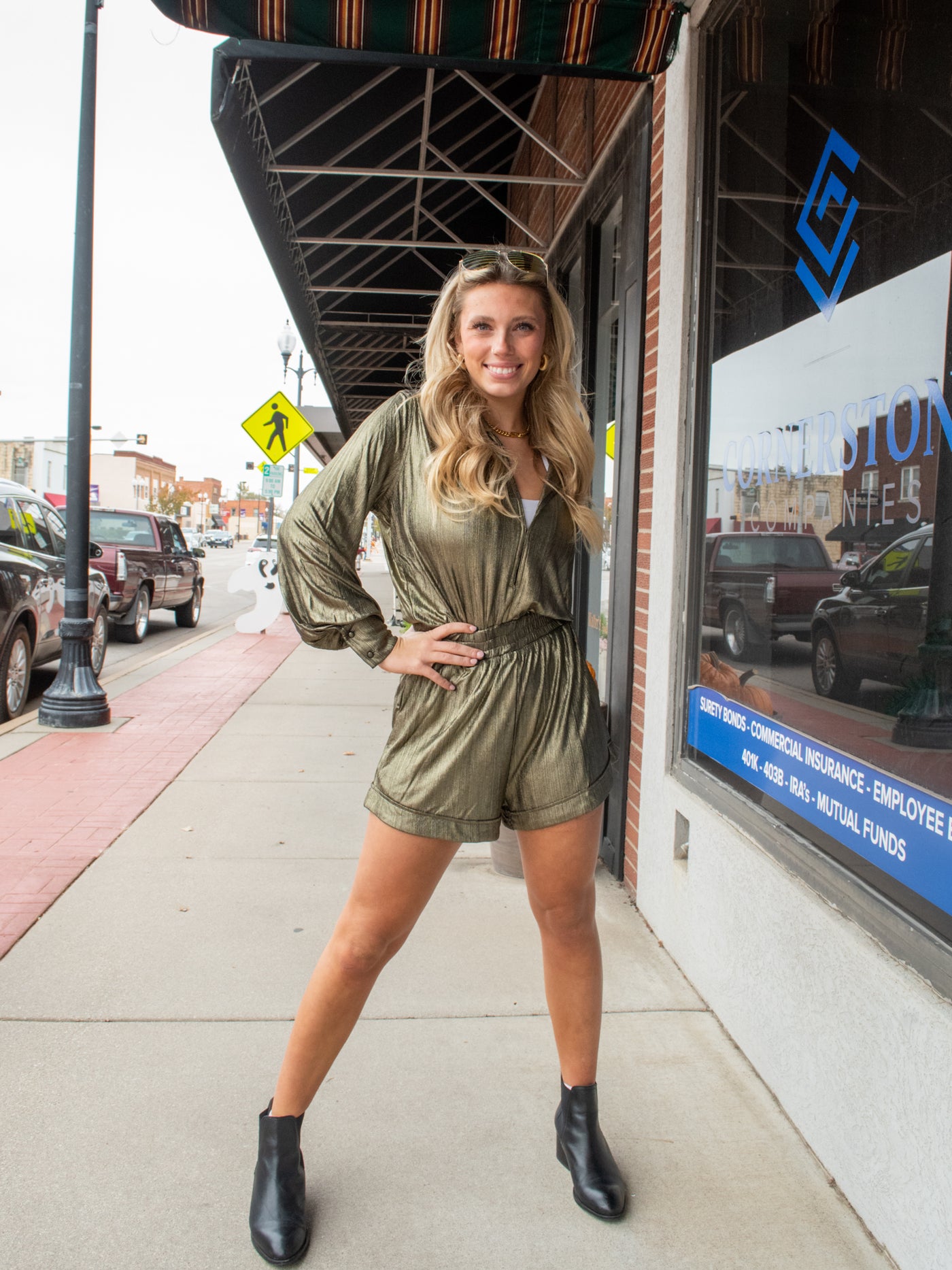 A model wearing a long sleeve, v-neck gold metallic romper with a waistband. She has it on with black booties and gold jewelry.