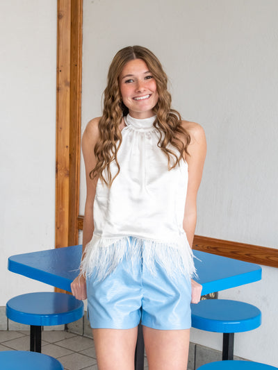 A model wearing a pair of light blue short with a white hatlerneck top with faux feather trim.