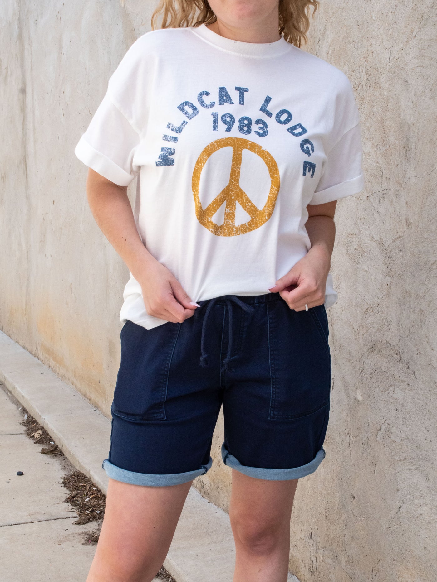 A model wearing a pair of dark wash drawstring sailor shorts with a white tee with a peach sign on it and white sneakers.