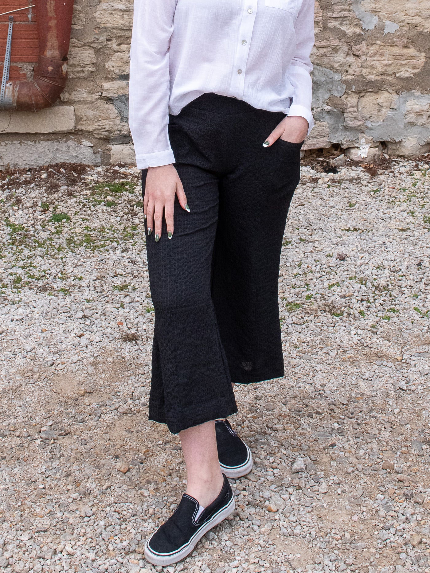 A model wearing a pair of wide leg crinkle ankle length pants with a white blouse and black loafers.