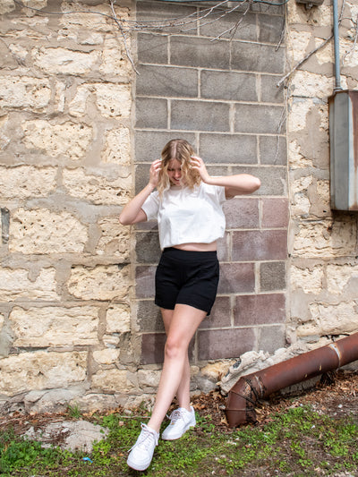 A model wearing a pair of black mid rise pull on crinkle material shorts with a white crewneck tee and white sneakers.