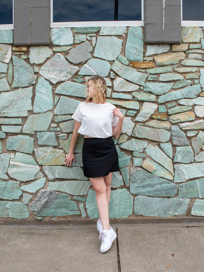 A model wearing a black crinkle material mini skort with with a white short sleeve crewneck tee and white sneakers.
