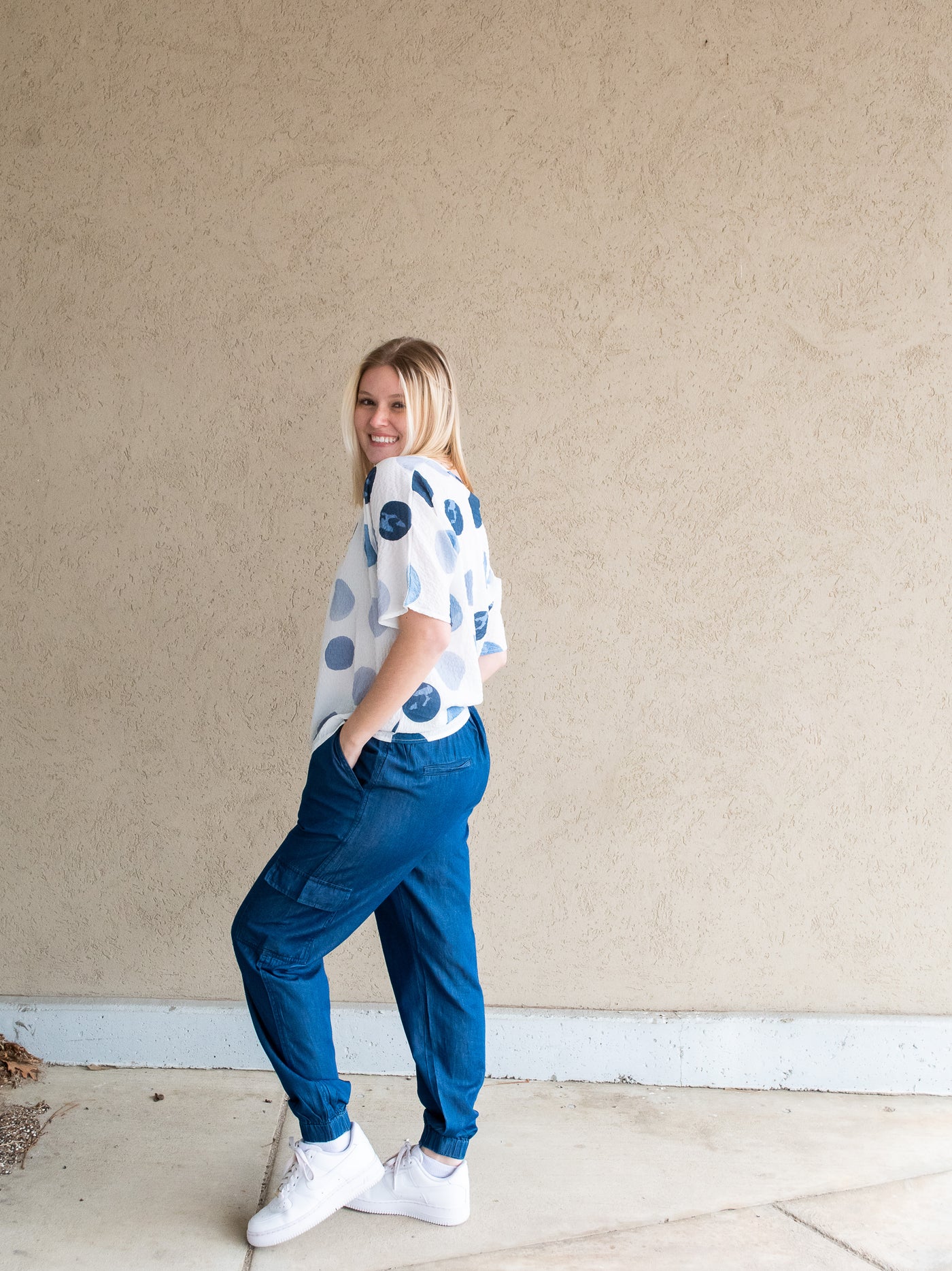 A model wearing a short sleeve white blouse with different shades of blue polka dots. She has it on with denim joggers and white sneakers.
