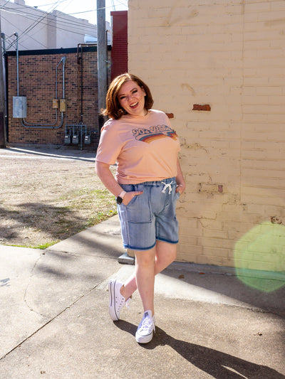 A model wearing a pair of light wash, drawstring waist shorts with a pink tee that says 90's baby on it. The model has it on with a pair of white sneakers.