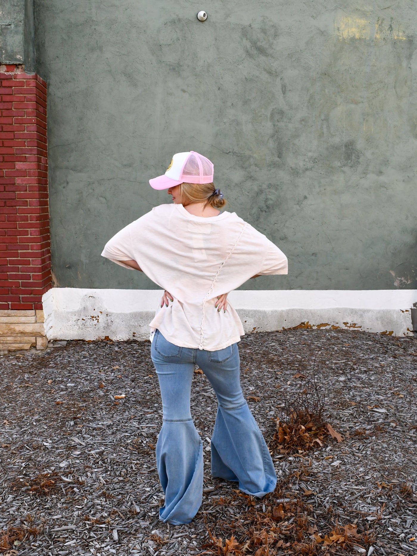 A model wearing a cream relaxed fit tee with braided details on the side. She has it paired with flare jeans and a trucker hat.