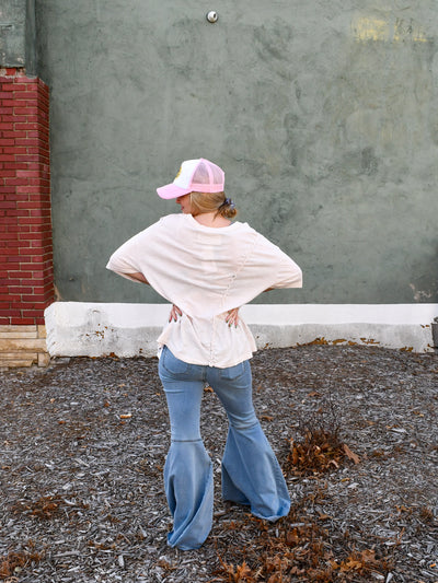 A model wearing a cream relaxed fit tee with braided details on the side. She has it paired with flare jeans and a trucker hat.