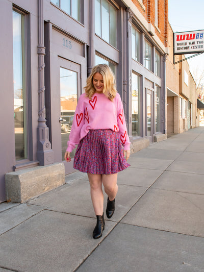 A model wearing a purple printed pleated skirt with a pink sweater with red hearts on it.