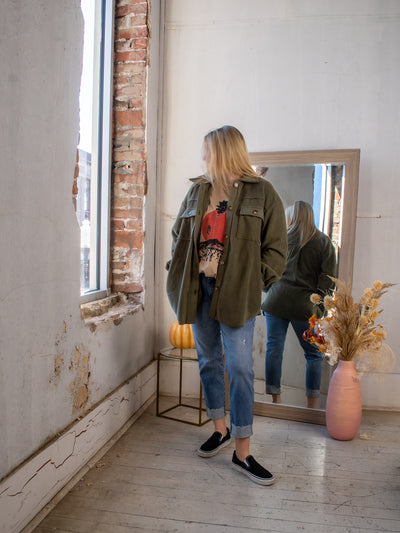 A model wearing an olive green shacket over a graphic tee and denim jeans with sneakers.