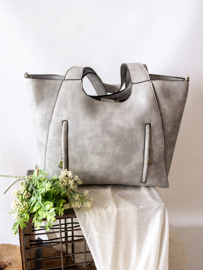 A grey tote bag with vertical zippers on the front tote straps and a snap flap closure. It as an optional strap you can attach and a small removable pouch that can snap into place on the inside.