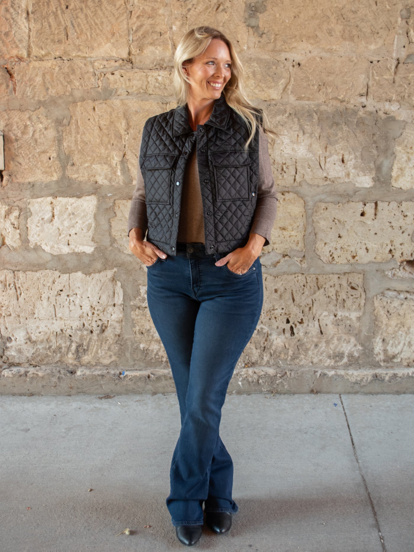 A model wearing a pair of dark wash bootcut jeans with a brown tee and a quilted black vest.
