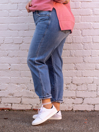 A model wearing a pair of ultra high rise slouch fit mom jeans. The jeans are rolled once at the bottom, have a zip fly closure with one button, a 5 pocket design. The model is wearing the jeans with white sneakers and a pink v neck sweatshirt that has "PRAY" on the front.