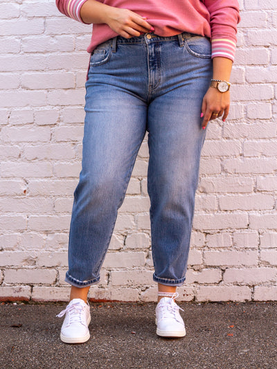 A model wearing a pair of ultra high rise slouch fit mom jeans. The jeans are rolled once at the bottom, have a zip fly closure with one button, a 5 pocket design. The model is wearing the jeans with white sneakers and a pink v neck sweatshirt that has "PRAY" on the front.