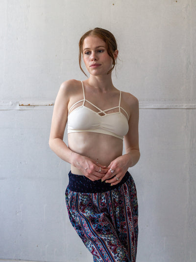 A model wearing an ivory bralette that is simple, stretchy material. It has two straps and a cage like design with two straps. Each strap is attached at the side seam and crosses over in front to attach to the other side about one inch from the small v neck. The main straps attach at the middle of each bra side and pull it up to form a peak.