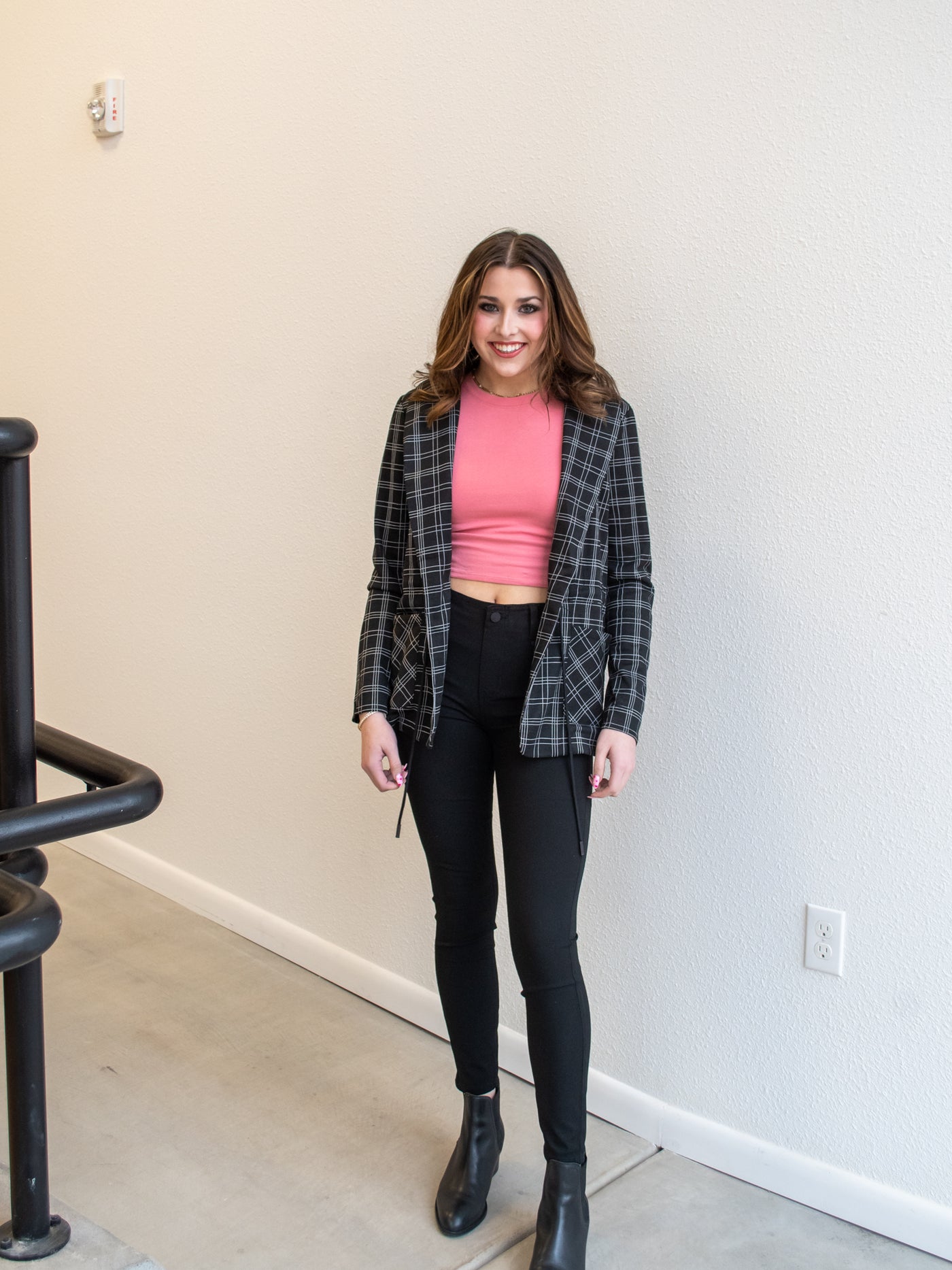 A model wearing a pair of black ankle length skinny pants with a pink tank, plaid blazer, and black booties.