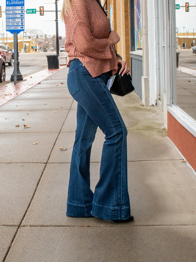 A model wearing a pair of high rise flare jeans with trim detailing. The model has them on with a blush crochet sweater over a matching tank and black booties.