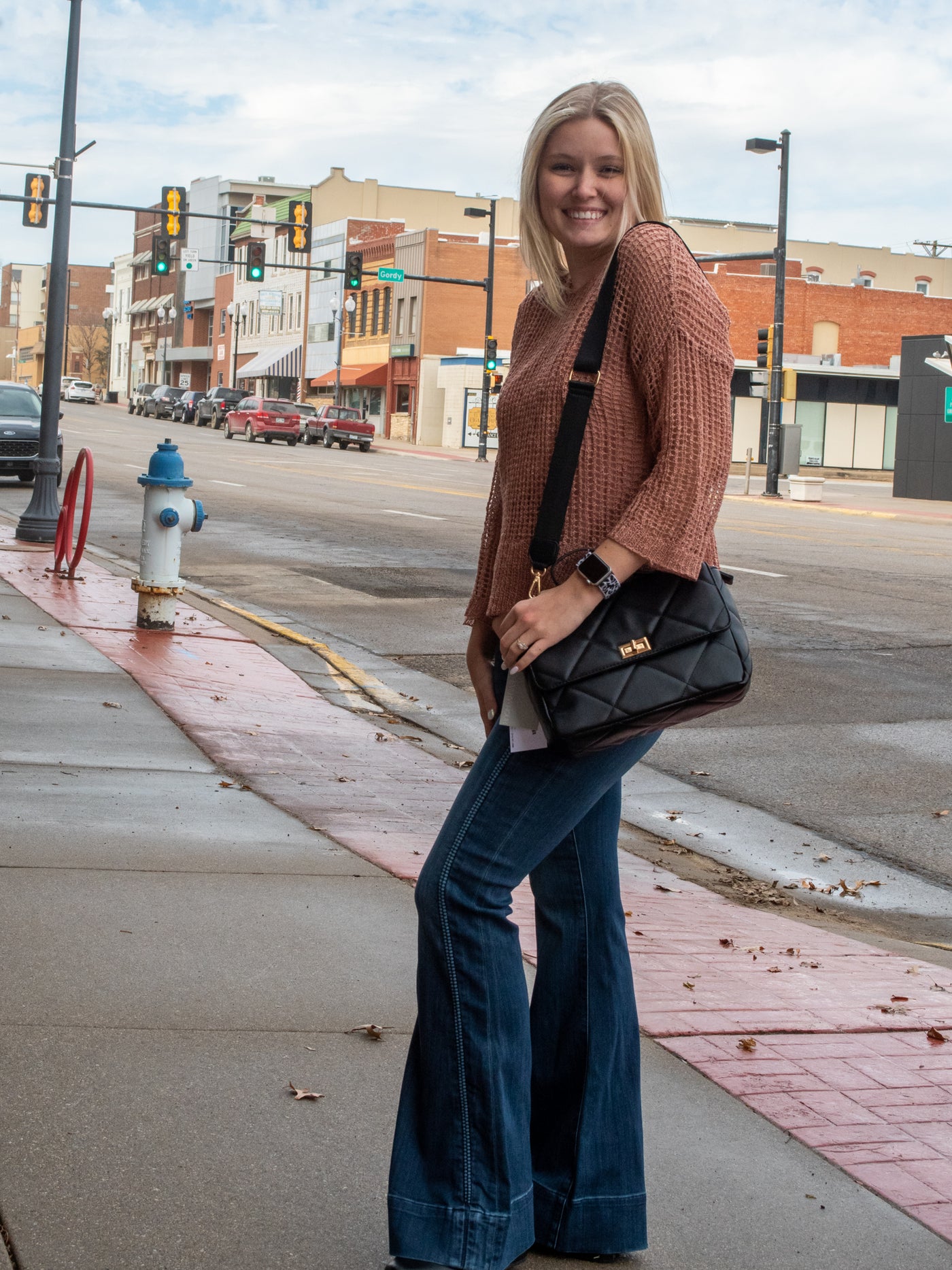 A model wearing a pair of high rise flare jeans with trim detailing. The model has them on with a blush crochet sweater over a matching tank and black booties.