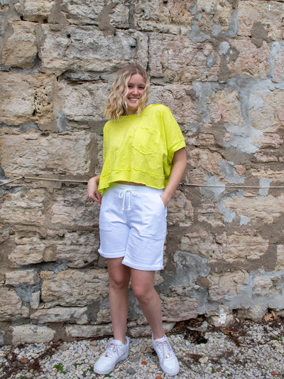 A model wearing a lime colored boxy slub jersey knit tee with raw edges and a front pocket. She has it on with white Bermuda shorts and white sneakers.
